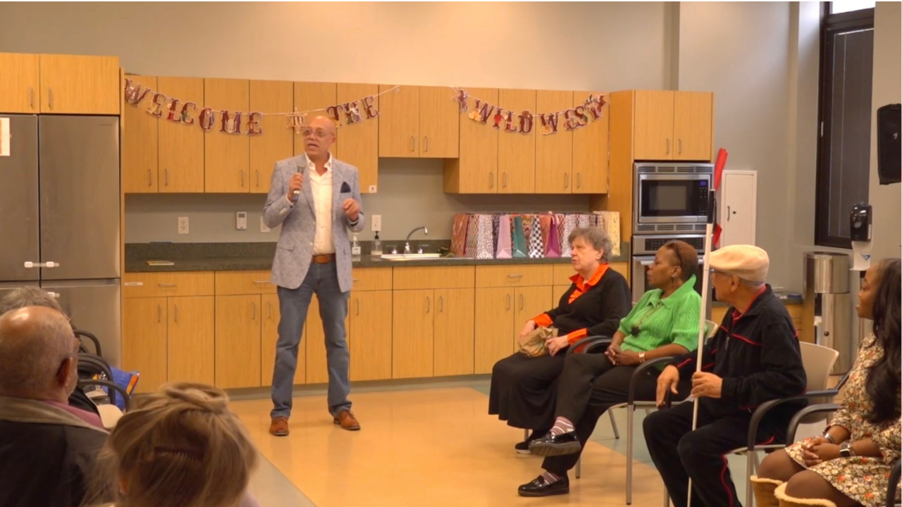 Todd Tolson stands at the front of a community gathering holding a handheld microphone as he addresses an audience. He wears clear-framed eyeglasses, a light blue patterned sport coat, a white dress shirt, jeans, and brown dress shoes. Behind him is a community kitchen with wood cabinets and a banner reading, "Welcome to the Wild West." Several attendees and panelist for the discussion listen attentively, including an older man holding a white cane, as Todd leads an engaging discussion in an accessible, conversational setting.
