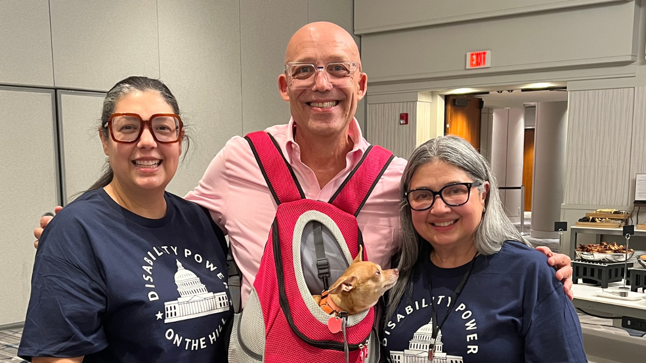 Todd Tolson smiles between two fellow volunteers during AAPD's Disability Power on the Hill event. He wears a light pink dress shirt and carries his small tan Chihuahua, Deja, in a bright pink front pet carrier. The two women, both wearing navy "Disability Power on the Hill" T-shirts and eyeglasses, stand with Todd, smiling toward the camera. The photo was taken in a conference hotel and reflects the spirit of advocacy, collaboration, and disability leadership.