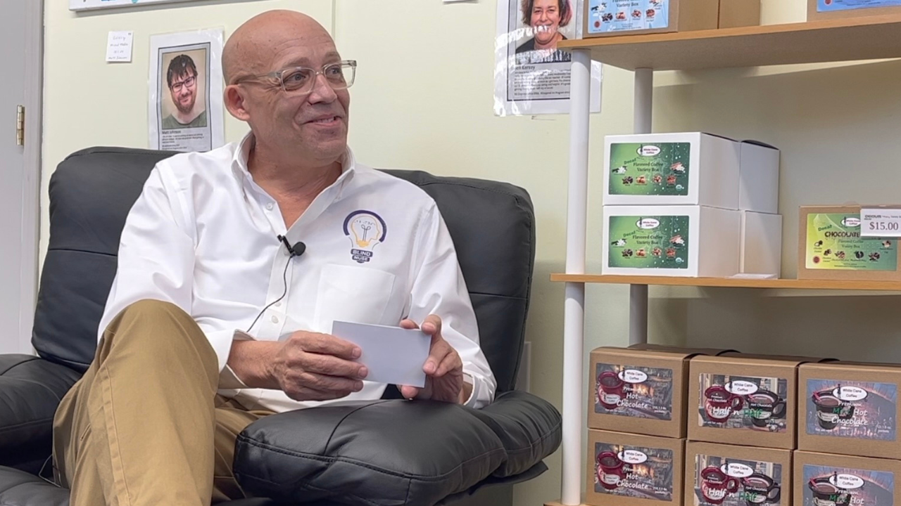 Todd Tolson sits in a comfortable chair during an on-camera interview, smiling as he holds a note card. He wears a white Blind Muse Foundation dress shirt with the organization's logo, khaki pants, and clear-framed eyeglasses. A lavalier microphone is clipped to his shirt. Shelves beside him display products from White Cane Coffee, creating the setting for a documentary interview highlighting entrepreneurship, accessibility, and community impact.