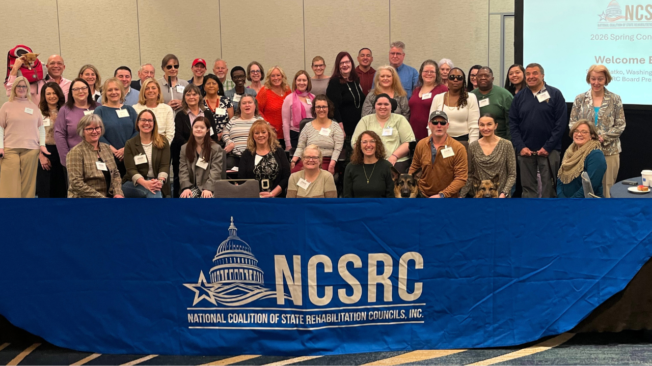 A large group of attendees from the National Coalition of State Rehabilitation Councils (NCSRC) Spring Conference pose together for a group photo behind a blue NCSRC banner displaying the organization's logo. The diverse group includes representatives from state rehabilitation councils across the country, several guide dogs, and participants with visible disabilities, reflecting the conference's commitment to collaboration, accessibility, and inclusion. Todd with his chihuahua Deja in a backpack on his shoulder is in the top left corner of the photo taken in a conference meeting room following the 2026 national gathering.