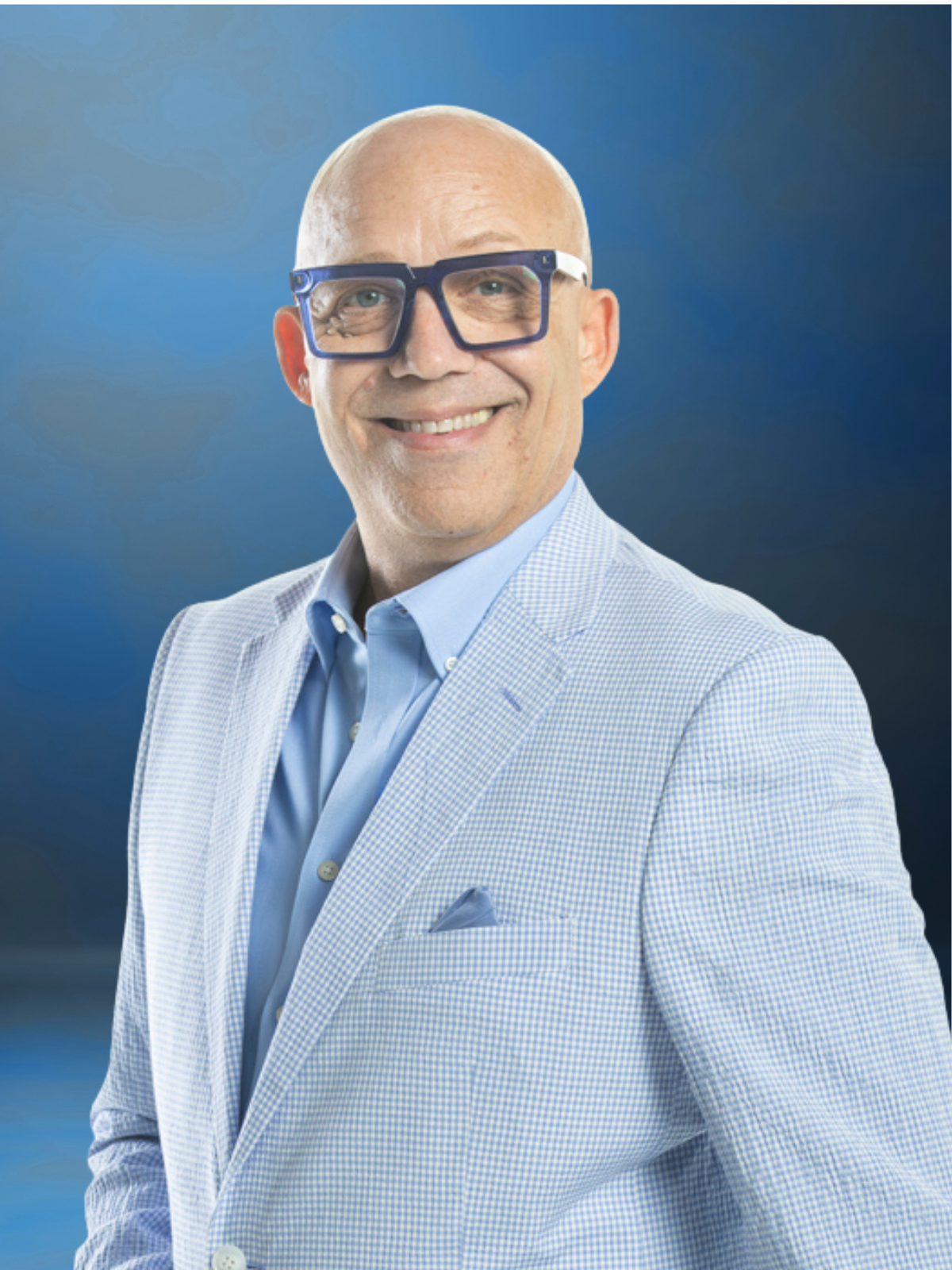 Todd Tolson smiles warmly while wearing blue-framed eyeglasses, a light blue dress shirt, and a light blue patterned sport coat with a matching pocket square. He stands against a soft blue gradient background, looking directly at the camera in a professional portrait that conveys confidence, warmth, and approachability.