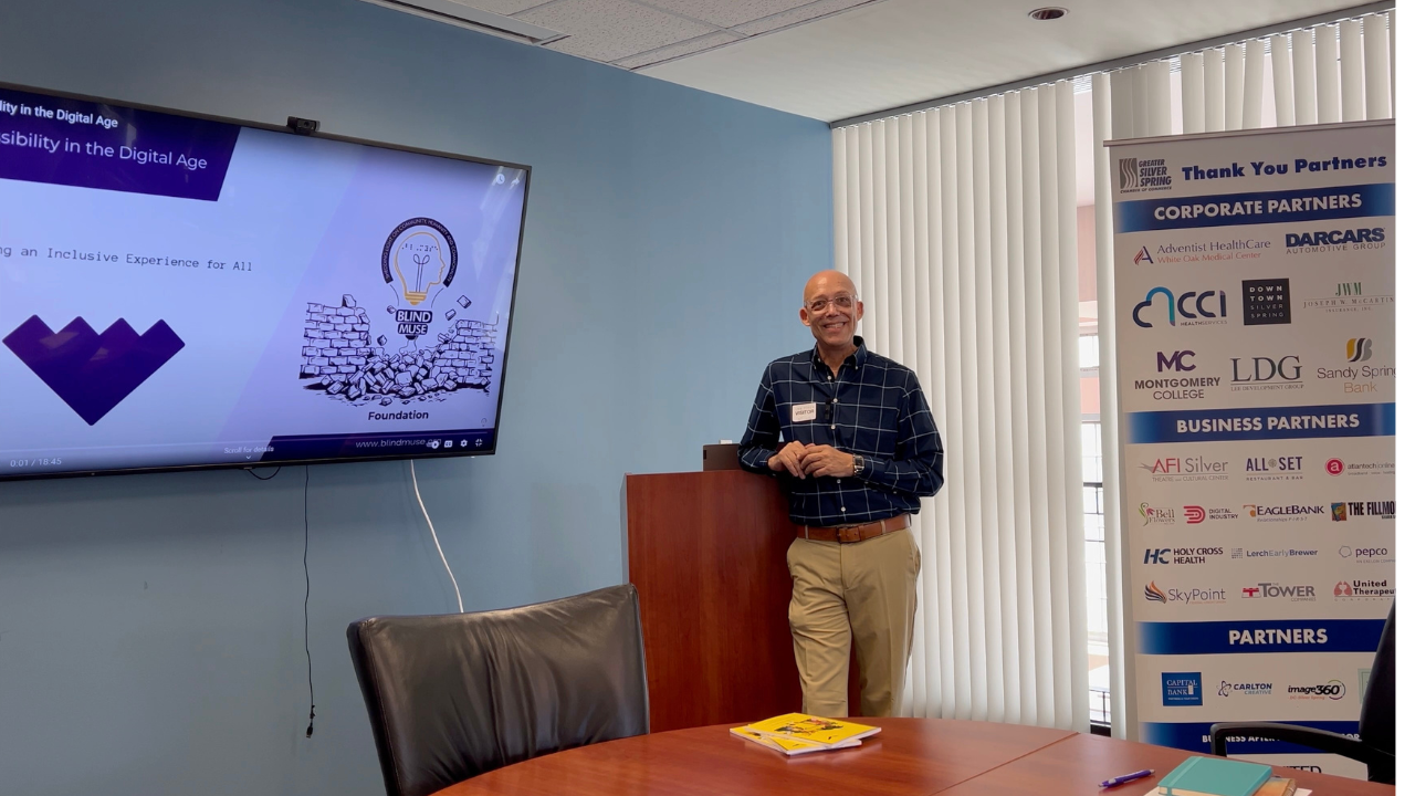 Todd Tolson stands beside a podium, smiling toward the camera, in a conference room at the Greater Silver Spring Chamber of Commerce. He wears a navy-blue checkered dress shirt, khaki pants, and a conference name badge. A large monitor displays a Blind Muse Foundation presentation on accessibility in the digital age, while a partner recognition banner featuring corporate and community organizations stands nearby. A conference table with meeting materials in the foreground reflects the professional setting where Todd is presenting on accessibility and inclusive communication.